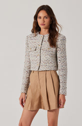 A woman is wearing a textured, multicolored woven cropped jacket with a round neckline and long sleeves. The jacket has decorative gold buttons and a front pocket on the left side. She pairs it with a beige, knee-length pleated skirt made of a lightweight fabric.
